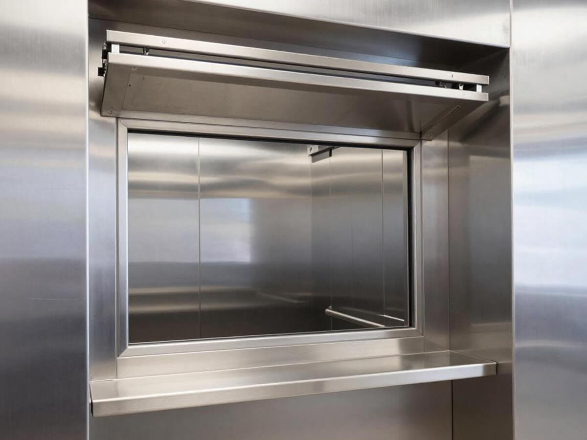 Open stainless steel dumbwaiter landing door showing the car interior set into a kitchen wall